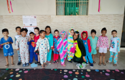 #paratodosverem: grupo de crianças posa sorridente em fila, vestindo pijamas coloridos e fantasias divertidas, como unicórnio e super-heróis. Ao centro, uma educadora de macacão verde também participa. O fundo é de parede clara com janela de grades e enfeites de papel pendurados. No chão, um tecido preto com bolinhas coloridas decora o espaço.