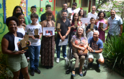 #paratodosverem: Um grupo misto de adultos e jovens, cerca de 16 pessoas, posa em pé e ajoelhado em um pátio externo da escola, cercado por folhagens. Vários membros do grupo seguram troféus, certificados ou pequenos objetos de premiação. Uma mulher está sentada em uma cadeira de rodas no centro. A parede atrás é pintada em cores vivas, incluindo verde e amarelo.