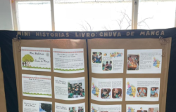 #paratodosverem: Um painel de exposição de papel pardo e preto com o título "MINI HISTÓRIAS LIVRO: CHUVA DE MANGA" está fixado em frente a uma janela. O painel exibe vários cartões de texto e fotos das crianças, organizados em colunas. As janelas, ao fundo, mostram uma paisagem externa com galhos de árvores secas.