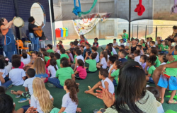 #paratodosverem: A imagem mostra um grupo grande de crianças sentadas no chão, assistindo a uma apresentação musical. Duas pessoas tocam instrumentos, uma com pandeiro e outra com violão, diante do público infantil. O evento ocorre em um espaço coberto, com parede azul e brinquedos coloridos ao fundo.