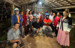 #paratodosverem: Um grupo de adultos posa em uma estrutura rústica de madeira e palha,. As pessoas aparecem sorrindo e reunidas.