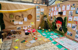 #paratodosverem: Canto expositivo sobre cultura indígena, com paredes forradas de papel pardo. Há artesanatos pendurados, cestos, colares e utensílios no chão sobre esteira. Ao centro, maquete de ocas. À direita, desenhos e fotos de alunos, além de um grande cartaz com grafismos.