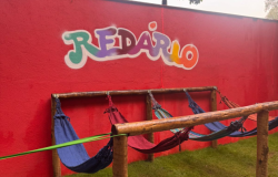 #paratodosverem: Área externa com uma parede vermelha onde está escrito “Redário” em letras coloridas. À frente, há uma estrutura de madeira com várias redes penduradas. O espaço é cercado por grama e árvores, criando um ambiente ao ar livre voltado para descanso e leitura.