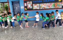 #paratodosverem: Um grupo de crianças, em fila, segura a cintura umas das outras e brinca em um pátio escolar. Elas usam uniformes verdes, azuis e brancos. Ao fundo, há um mural colorido com pinturas feitas pelas próprias crianças.