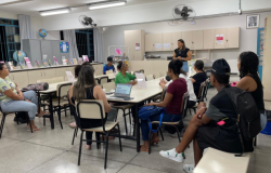 #paratodosverem: Em uma biblioteca escolar, um grupo de alunos da EJA está sentado ao redor de mesas organizadas em formato de reunião. Uma mulher está em pé à frente, falando e conduzindo a atividade. Sobre as mesas, há papéis e um notebook. Ao fundo, estantes com livros e materiais organizados indicam o espaço da biblioteca.