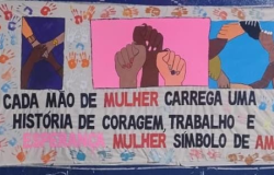 #paratodosverem: Painel colorido com ilustrações de mãos dadas e erguidas, representando união e diversidade. Ao centro, há a frase: “Cada mão de mulher carrega uma história de coragem, trabalho e esperança. Mulher símbolo de amor.” O fundo é decorado com várias marcas de mãos coloridas, sugerindo participação coletiva.