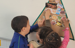 #paratodosverem: Três crianças participam de uma atividade em sala de aula, organizando figuras de alimentos em uma pirâmide alimentar fixada na parede. Enquanto colam as imagens, aprendem sobre os grupos alimentares e a importância de uma alimentação saudável de forma colaborativa e lúdica 