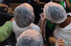 #paratodosverem: Grupo de estudantes reunidos ao redor de uma lixeira. As crianças usam toucas descartáveis e observam a atividade. A ação acontece na área da cozinha da escola e integra o projeto de conscientização sobre o desperdício de alimentos. 