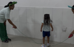 #paratodosverem: Em sala de azulejos brancos, duas adultas seguram uma corda no chão. À esquerda, mulher de calça verde usa tiara de folhas verdes. No centro, criança de costas com shorts azuis e tênis de luzes azuis acesas está sobre a corda. À direita, outra adulta usa a mesma tiara.