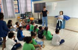 #paratodosverem: Sala de aula com vários alunos sentados no chão, enquanto uma colega em pé fala à frente da turma, ao lado de uma mulher. A sala tem um quadro branco, um televisor e janelas com cortinas brancas. As crianças vestem uniformes escolares da Prefeitura de Belo Horizonte