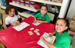 #paratodosverem: três estudantes sentadas em uma mesa vermelha manuseiam álbuns e figurinhas soltas de futebol. Duas vestem uniformes verdes e uma uniforme branco, todas sorrindo. Ao fundo, prateleiras com caixas organizadoras rotuladas como "CLÉO", "COLETIVO" e "ANA LUIZA", e uma maleta vermelha de primeiros socorros. O chão é de ladrilhos amarelos.