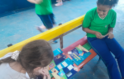 #paratodosverem: Duas alunas sentadas em banco colorido com encosto amarelo organizam um "Álbum de Figurinhas Literárias" aberto entre elas. À direita, uma estudante de camiseta verde e calça azul manuseia figurinhas. À esquerda, em primeiro plano, outra estudante observa o álbum e cola um adesivo. O chão da área externa é azul. Ao fundo, outra criança em movimento passa correndo.