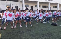#paratodosverem: Grupo de crianças faz uma apresentação em campo de futebol. Elas usam camisetas brancas com desenhos de coração rosa e seguram fitas brancas nas mãos durante dança