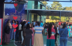 #paratodosverem: Foto mostra um grupo de adultos reunidos na área externa da EMEI Belmonte, em um espaço coberto. As pessoas conversam e circulam próximas à quadra. O ambiente está decorado com corações vermelhos pendurados e balões. Ao fundo, aparecem paredes coloridas da escola, árvores e parte da cobertura metálica. Uma das participantes veste uma blusa listrada.