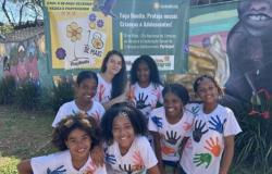#paratodosverem: Fora da escola, sete estudantes (todas meninas) posam sorrindo em frente a um painel da campanha Maio Laranja, com mensagens de proteção à infância e adolescência. Elas vestem camisetas brancas com pinturas coloridas de mãos.