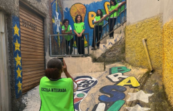 #PARATODOSVEREM: Quatro estudantes com coletes verdes posam em uma escadaria cercada por muros grafitados. Em primeiro plano, outra aluna fotografa o grupo com um celular. O local possui pinturas coloridas no chão e nas paredes, além de corrimãos e construções ao redor. 