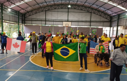 #paratodosverem: grupo de pessoas reunido em uma quadra poliesportiva coberta celebrando com entusiasmo. Elas seguram bandeiras de diferentes países, como Brasil, Canadá, Estados Unidos e México, além de adereços verdes e amarelos
