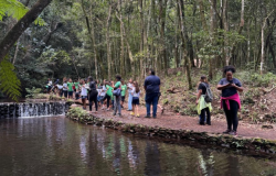 #PARATODOSVEREM: Grupo de estudantes caminha por uma trilha ao lado de um curso d’água em uma área de mata. Alguns alunos observam o ambiente, enquanto outros utilizam celulares. Ao fundo, há árvores altas e uma pequena queda d’água cercada por pedras. 