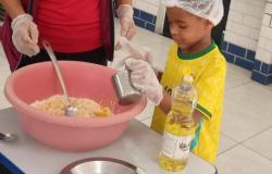 #paratodosverem: Menino usando touca e luvas descartáveis durante uma atividade culinária. Ao lado de uma educadora, ela segura um copo ao lado de uma mesa com uma tigela rosa contendo ingredientes e uma garrafa de óleo. 