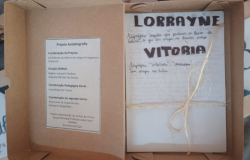 #paratodosverem: Uma caixa de papelão aberta com um livro artesanal amarrado com barbante, trazendo o nome “Lorrayne Vitória” na capa e informações sobre um projeto escolar de autobiografia.