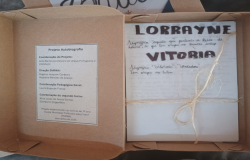 #paratodosverem: Uma caixa de papelão aberta com um livro artesanal amarrado com barbante, trazendo o nome “Lorrayne Vitória” na capa e informações sobre um projeto escolar de autobiografia