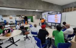 #paratodosverem: Em uma sala com mesas e cadeiras, homens e mulheres alunos da EJA estão sentados acompanhando uma palestra. Ao fundo, uma mulher de camiseta laranja, do Maio Laranja, apresenta conteúdos em uma tela digital sobre prevenção à violência e proteção de crianças e adolescentes. O ambiente possui estantes, quadro branco e materiais educativos. 
