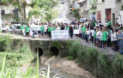 #paratodosverem: Grande grupo de estudantes e educadores reunidos em uma rua, ao lado de um córrego canalizado, durante uma caminhada. Os participantes seguram faixas e cartazes, com destaque para o banner “2ª Caminhada das Águas”, da Escola Municipal Rui da Costa Val, datado de 24-03-2026. Ao redor, há casas, vegetação e uma ponte com duas manilhas por onde passa a água.