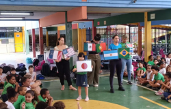 #PARATODOSVEREM: No pátio coberto da escola, dezenas de crianças estão sentadas no chão em semicírculo assistindo à atividade. Ao centro, uma aluna e quatro educadoras seguram cartazes com bandeiras de países como Japão, França, EUA, México, Portugal, Argentina e Brasil. O ambiente escolar é colorido, com colunas em tons de azul, amarelo e vermelho, além de mochilas organizadas ao fundo. 