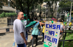 #paratodosverem: Um grupo de pessoas caminha em um desfile ao ar livre. Elas usam roupas coloridas e carregam um cartaz com a frase “EMSA na folia”, decorado com corações e a bandeira do Brasil. Ao fundo, aparecem prédios.