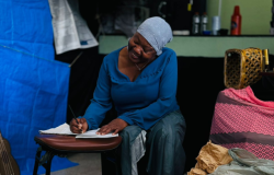 #paratodosverem: Mulher negra, sentada em um banco, usa um lenço na cabeça e roupa azul. Ela escreve concentrada em um caderno apoiado em um banquinho. O espaço ao redor tem objetos como sacolas, uma cadeira e um violão ao fundo, sugerindo um cenário de teatro que remete ao cotidiano retratado na obra.