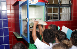 #paratodosverem: Estudantes de costas em uniformes brancos e verdes cercam uma caixa de madeira azul e verde em parede de azulejos vermelhos e azuis. A caixa aberta tem a placa "Emei Minaslândia". Várias mãos pequenas colocam os livros na caixa. Há uma janela de ferro no fundo.