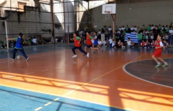 #paratodosverem: Em quadra poliesportiva bicolor (laranja e azul), acontece uma partida de futsal. Quatro estudantes em movimento vestem coletes vermelhos e azuis sobre seus uniformes. O estudante da esquerda, vestindo azul, conduz a bola. No fundo, há uma grade de proteção e uma arquibancada com muitos estudantes sentados assistindo. Uma cesta de basquete e uma trave de gol estão visíveis.