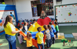 #paratodosverem: Em uma área externa da escola, crianças participam de uma brincadeira com um grande pano com vários retalhos nas cores vermelho, amarelo, azul e laranja. Duas educadoras acompanham a atividade. Algumas crianças usam camisas de times de futebol. Ao fundo, há brinquedos infantis e murais decorados na parede. 