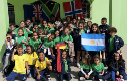 #paratodosverem: Estudantes segurando bandeiras de diferentes países. As crianças sorriem para a foto e usam uniformes nas cores verde, azul e amarelo. Ao fundo, há bandeiras penduradas na parede.