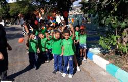 #paratodosverem: Grupo de crianças com uniformes escolares (camisas verdes e calças azuis) caminham acompanhados por adultos. Os alunos seguram cataventos coloridos e balões vermelhos, enquanto olham para a câmera. 