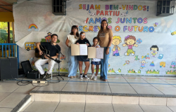 #paratodosverem: A foto mostra duas crianças ao centro segurando um cartaz aberto, acompanhadas por duas mulheres adultas ao lado delas. À esquerda, um homem toca violão sentado. Todos estão em frente a um painel com a frase “Sejam bem-vindos! Vamos juntos escrever o futuro.”