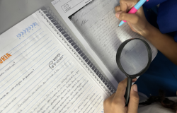 #paratodosverem: Imagem vista de cima mostra as mãos de um estudante escrevendo em um caderno enquanto segura uma lupa sobre um documento antigo impresso. Ao lado, há outro caderno com anotações feitas à mão. A cena sugere uma atividade escolar de análise de fontes históricas.