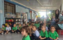 #paratodosverem: Em espaço coberto da escola, crianças pequenas uniformizadas se sentam no chão e observam atentamente uma atividade fora do enquadramento. Elas vestem uniformes nas cores verde, branco e azul. Educadoras e alguns responsáveis acompanham o momento também sentados.