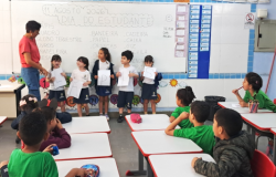 #paratodosverem: Em uma sala de aula, cinco alunos de uniforme estão alinhados em frente ao quadro branco segurando papéis com as letras L, Á, P, I, S, orientados por uma professora de blusa vermelha. No quadro está escrito "11 AGOSTO 2026 DIA DO ESTUDANTE". Em primeiro plano, outros alunos sentados em carteiras brancas com bordas vermelhas observam a apresentação. Acima do quadro, há um alfabeto.