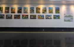 #paratodosverem: Exposição com 18 pinturas coloridas de paisagens e construções históricas expostas em uma parede branca da galeria. As obras retratam cenas de Ouro Preto, com igrejas, ruas e casas em diferentes estilos e cores. Ao lado, há um painel informativo com o título “Ouro Preto: Nosso Patrimônio”.