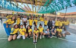 #paratodosverem: Em uma quadra coberta e decorada com bandeirinhas do Brasil e das cores verde, amarelo e azul, estudantes e educadores posam para fotografia em grupo. A maioria veste camisetas amarelas inspiradas no uniforme da Seleção Brasileira e usa medalhas no pescoço. Algumas crianças estão sentadas à frente, enquanto outras permanecem em pé ao lado dos professores.