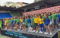 #paratodosverem: Um grupo com várias crianças se apresenta em um palco preto montado no pátio de uma escola. Elas vestem camisas verdes, azuis ou amarelas e shorts jeans. À frente do palco, está escrito "FESTA DA FAMÍLIA 2026 FAMÍLIA: NOSSO TIME CAMPEÃO". Ao fundo, há um painel decorado com corações vermelhos e brancos.