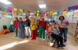 #paratodosverem: Sete mulheres posam sorridentes sob bandeirinhas coloridas de Festa Junina em um salão iluminado. Elas vestem trajes casuais e xadrez, segurando plaquinhas divertidas com frases como "MODO ARRAIÁ ATIVADO". Entre elas, uma mulher está em uma cadeira de rodas. Ao fundo, há mesas compridas organizadas para a celebração e uma caixa de som sobre uma pequena mesa laranja à esquerda.