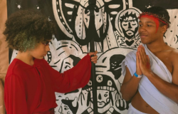 #paratodosverem: Dois jovens atuam em uma cena teatral. À esquerda, um jovem de cabelos cacheados e blusa vermelha interpreta o Diabo, segurando um tridente preto e olhando fixamente para o outro. À direita, um jovem de túnica branca, coroado com espinhos e mãos postas em oração, representa Jesus. Ao fundo, um painel ilustrado em estilo cordel em preto e branco exibe figuras sacras e nordestinas.