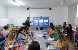 #paratodosverem: Educador apresenta conteúdo sobre IA em sala de aula, usando uma tela interativa. Os demais professores, sentados ao redor de uma mesa com laptops e cadernos, acompanham atentos e participam da discussão em um ambiente colaborativo de aprendizagem.