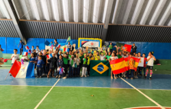 #paratodosverem: Um grande grupo de crianças posa reunido em uma quadra poliesportiva de piso verde e azul, sob teto metálico. Elas seguram bandeiras de diferentes países, como Brasil, França, Espanha e Portugal, além de erguerem troféus. Ao fundo, há uma parede azul e um painel emoldurado por balões amarelos e azuis, caracterizando uma gincana escolar.