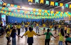 #paratodosverem: Em quadra coberta, estudantes participam de uma atividade, vestidos com roupas nas cores verde e amarelo. No centro, eles formam uma roda de mãos dadas. O ambiente está decorado com bandeirinhas juninas também das cores verde e amarelo no teto, enquanto outros alunos acompanham a programação ao redor da quadra. 