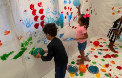 #paratodosverem: A imagem mostra três crianças brincando em um espaço coberto por papel branco no chão e paredes. Elas seguram balões mergulhados em tinta colorida e os usam para pintar o papel, criando marcas de mãos e pintura. O chão está cheio de pratos metálicos com tinta de várias cores, como rosa, azul e amarelo.