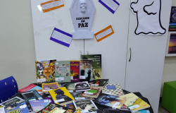 #paratodosverem: A imagem mostra uma exposição temática de livros intitulada “Beco do Arrepio”, montada em um ambiente escolar. A decoração inclui elementos que remetem ao Halloween, como uma lápide com os dizeres “Descanse em Paz”, teias de aranha, aranhas de brinquedo e a figura de um fantasma na parede. Sobre a mesa, há uma grande variedade de livros de mistério, terror e suspense.