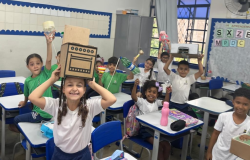 #paratodosverem: Em uma sala de aula iluminada, um grupo de crianças está animado em suas carteiras, segurando objetos reciclados acima da cabeça. Uma menina no centro ri, usando uma caixa de papelão desenhada como fogão.